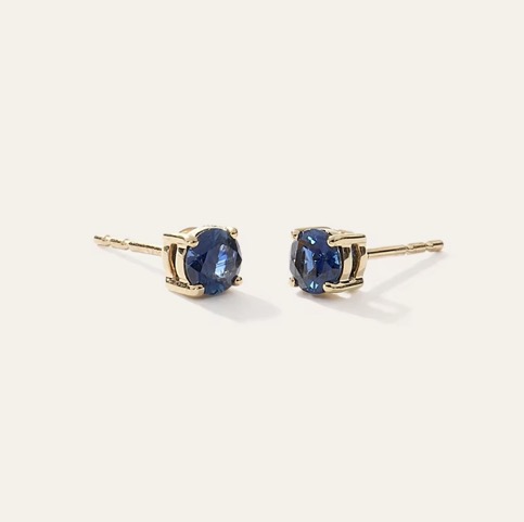 Pair of 14k gold solitaire stud earrings with round blue sapphires in four-prong settings on a cream background.